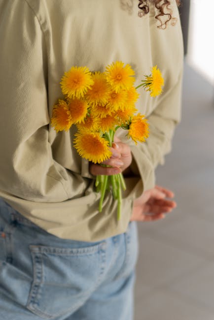 A person wearing a beige shirt and light blue jeans is holding a bouquet of bright yellow flowers, including daisies and pom-pom chrysanthemums, wrapped casually in the person's hand. The flowers appe