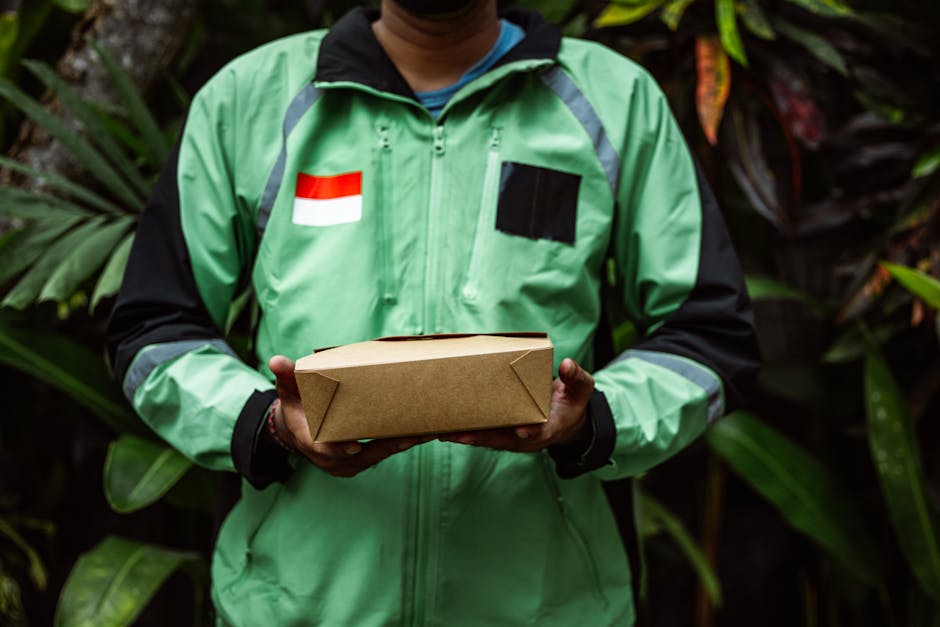 A delivery person is holding a rectangular, brown cardboard flower delivery box with both hands, positioned in front of their torso. They are wearing a bright green and black jacket featuring a small 