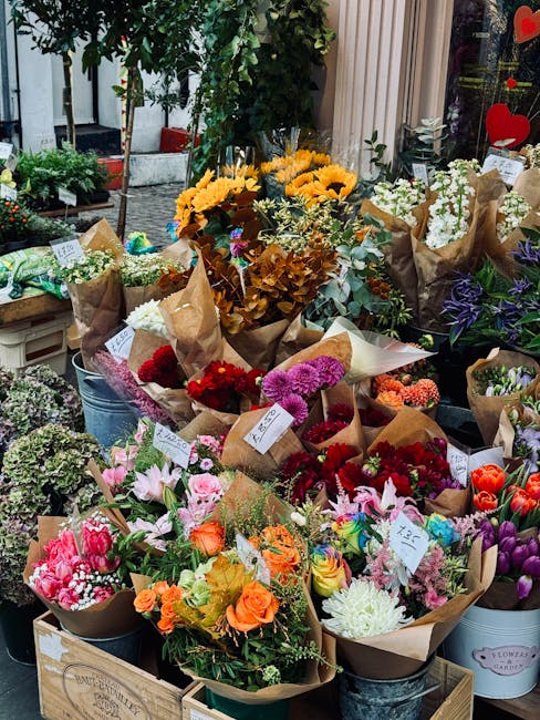 A vibrant display of freshly gathered flowers arranged in bouquets and potted plants at a flower shop. The arrangement includes yellow sunflowers, pink and white lilies, orange roses, red and purple c