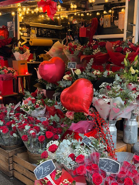An indoor floral display featuring a variety of fresh red roses, white baby's breath, and green foliage arranged in wicker baskets and vases. The bouquets are wrapped in clear and colored paper, with 