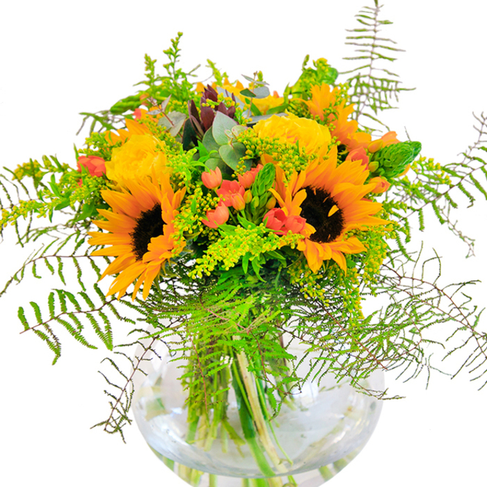Pure sunlit energy radiates from this florist-crafted bouquet-a tribute to Herne Hill's bustling street culture and its verdant, welcoming parks. Large golden sunflowers lead the arrangement, their faces wide and open, echoing the bright floral displays at Herne Hill Market on a cheerful weekend. Illuminated alongside are spiralling yellow roses, petals unfurling with elegant warmth, infusing the ensemble with a gentle fragrance suggestive of nearby woodlands around Ruskin Park. Scarlet Hypericum berries nestle amid a tapestry of greens, their glossy skins catching the eye and recalling the ripened fruit sold on Norwood Road. The composition brims with textural complexity: feathery ferns and solidago spill outwards, lending lightness, while subtle blue-green eucalyptus leaves weave throughout. All are carefully anchored in a globe-shaped clear vase, its water crystal and the stems fresh-cut for enduring vibrance. Each element is chosen to celebrate life's happy milestones in Herne Hill-be it a local birthday, family gathering, or simply bringing the outside in after a sunny walk. The bouquet's sensory notes-colour, scent, and touch-offer an exuberant, joyful mood ideal for lighting up any SE24 space.
