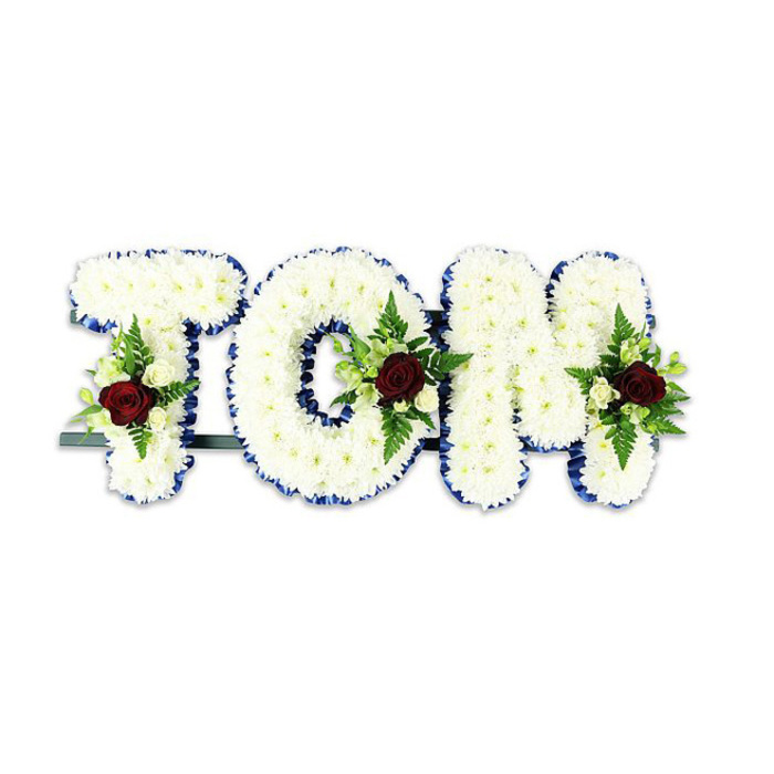 Presented against a simple white background, this bespoke sympathy piece spells TOM in three-dimensional lettering built from tightly grouped white chrysanthemums, creating a soft, voluminous surface that reads as both bold and tender. Each letter's outline is finished with a striking royal blue ribbon, the colour recalling the deep evening sky over Brockwell Park, and the ribbon's clean edge brings a precise geometry to the otherwise cloud-like texture of the blooms. Placed within each letter is a modest, symmetrical spray: a solitary deep red rose anchors the arrangement, chosen for its velvety depth and association with lasting affection; it is surrounded by small, creamy white spray roses and tiny buds, their subtle scent mingling with the green freshness of fern fronds and glossy foliage. The composition balances formality and comfort - the white chrysanthemums convey purity and quiet support, while the red roses speak to enduring memory. A slim dark metal support bar is visible behind the letters, confirming the tribute is built to stand upright at a service or graveside, crafted for visibility and reverence. The overall atmosphere in the image feels like a calm, respectful hush - appropriate for a chapel near Herne Hill Road or a small ceremony close to the station - where every petal placement reads as an act of care by the florist's hand. This arrangement exemplifies a personalised memorial option, a quiet, tactile way to say goodbye while celebrating a life remembered lovingly by family and friends.