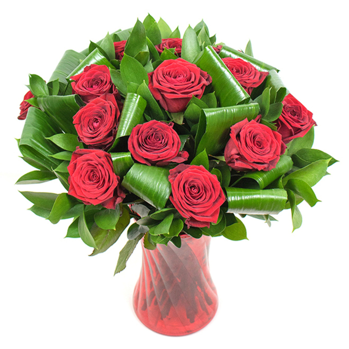 In this photograph a dense cluster of deep crimson roses commands attention, each bloom opened fully so its velvety petals spiral from a tight centre into soft, layered edges that catch the light in subtle variations of red. Surrounding and lifting the roses is an abundant mix of dark green foliage: delicate blade-like leaves that build volume and larger, glossy leaves folded and rolled into elegant cylinders and arcs which cradle the flowers and introduce sculptural movement. The contrast between the plush matte of the rose petals and the polished sheen of the greens is heightened by gentle, even illumination that reveals veins, curvature, and the tactile quality of every surface. The stems are contained within a tall, narrow, translucent ruby-red glass vase with a faintly ribbed, twisted pattern; the vase's slender waist and flared top create a poised silhouette that complements the arrangement's vertical rhythm and accentuates the intensity of the reds. Placed on a stark white background, the bouquet reads as an isolated statement of romantic refinement - the kind of piece a florist in Herne Hill would recommend for a classic anniversary gift or a thoughtful Valentine sent to someone living near Herne Hill station or enjoying a picnic in Brockwell Park. The image suggests the soft perfume of rose, a careful hand in leaf placement, and a design that marries traditional symbolism with contemporary styling. It speaks to considered craftsmanship, freshness, and an emotional warmth suitable for intimate celebrations or as a striking focal point in a calm, modern interior.