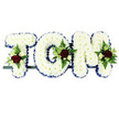 A carefully constructed floral name tribute spells TOM in bold block letters, the kind of bespoke funeral arrangement often chosen for services around Herne Hill and Brockwell Park. Each letter is entirely filled with pristine white chrysanthemums, their small blooms tightly clustered to create a dense, velvety texture and an even, snow-like surface. Tracing the outer edges, a royal blue satin ribbon provides a smooth, lustrous frame, neatly folded and pinned to follow every corner and curve, giving the design a polished and respectful finish. At strategic points on the letters-near the top left of the 'T', the curve of the 'O', and the upper right of the 'M'-compact floral sprays introduce rich colour and texture: deep red roses fully open at the centre, surrounded by dainty white or cream spray roses and slim, white, freesia-style flowers. These are anchored in small clusters of fresh foliage, including feathery fern fronds and glossy leaves, which soften the edges and add a natural, breathing quality to the piece. The letters are lined up on a narrow dark frame, designed for secure placement at a funeral, perhaps resting beside the coffin in a chapel near Railton Road or displayed at a crematorium service for family and friends to see. The plain white background in the image heightens the contrast between the pure white chrysanthemums, the deep red roses, and the royal blue ribbon. Altogether, the tribute conveys a sense of purity, devotion, and quiet strength, honouring Tom's name with flowers that speak gently of love and remembrance.