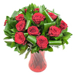A richly detailed, rounded bouquet of a dozen or more deep crimson roses sits like a jewel in this image, each bloom open to reveal velvety, tightly spiralled petals that soften outward in layers of plush red. The roses are woven through with generous, glossy dark green foliage - fine-leaved sprigs that build a dense backdrop and broader, architectural leaves rolled and curled into sculptural loops that cradle the rose heads like evergreen ribbons. Those folded, emerald leaves catch the light with a satin sheen, their smooth surfaces contrasting with the delicate, matte texture of the rose petals. The stems rest in a tall, slender, translucent ruby-red glass vase whose faint ribbed or twisted pattern creates vertical lines that catch highlights and echo the passionate tones of the flowers. Soft, even lighting from the front reveals subtle petal ridges and the cool shine of the greens while a crisp white background isolates the arrangement so every scarlet bloom and curled leaf reads clearly. As a local florist might describe it to a customer from Herne Hill or someone strolling past Brockwell Park, the bouquet evokes classic romance and considered contemporary design - the scent almost perceptible, a warm, heady perfume that suggests anniversary dinners, Valentine's surprises, or an elegant table centrepiece at a small, intimate gathering. The composition balances traditional symbolism and modern styling: lush, abundant roses for timeless devotion, trimmed and shaped foliage for structure and movement. This view communicates freshness, meticulous handwork, and the kind of visual presence that lifts a room or marks a cherished occasion with confident, sophisticated colour.