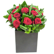 In this elegantly modern arrangement, passion and refined structure sit comfortably side by side. A tight cluster of velvety crimson roses forms the luminous centre, each bloom opening in soft spirals whose petal edges catch the bright, clear studio light and glow with a velvet sheen. Surrounding the roses, broad, glossy green leaves have been artfully curled and folded into rhythmic loops, their sculptural shapes adding movement and depth while creating a crisp contrast to the softness of the blooms. The bouquet is set in a matte black, square cube that anchors the composition with a contemporary, geometric calm; its flat surface absorbs light so the roses appear even more vivid above. If you linger, there is a suggestion of scent - the classic heady sweetness of rose balanced by clean, almost tropical green notes from the foliage, like a whisper of fresh-cut leaves after rain.
As a local florist familiar with Herne Hill, I imagine this piece gracing a contemporary flat near Brockwell Park or standing proudly on a café table along Railton Road, where architecture and greenery meet. The lighting feels modern and honest, revealing glossy highlights on each leaf and the delicate velour of each petal. Texturally, the interplay between soft, yielding petals and the firmer, waxy leaves invites touch without overwhelming - a design that reads as both dramatic and composed.
This arrangement is intended as a mindful, romantic gesture: perfect for anniversaries, intimate celebrations, or for bringing quiet theatre to an urban living room. Its confident simplicity and crafted detail make it a lasting floral statement for any occasion in and around Herne Hill.
